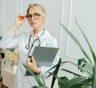 Confident female doctor wearing eyeglasses and holding a clipboard in an indoor office setting.